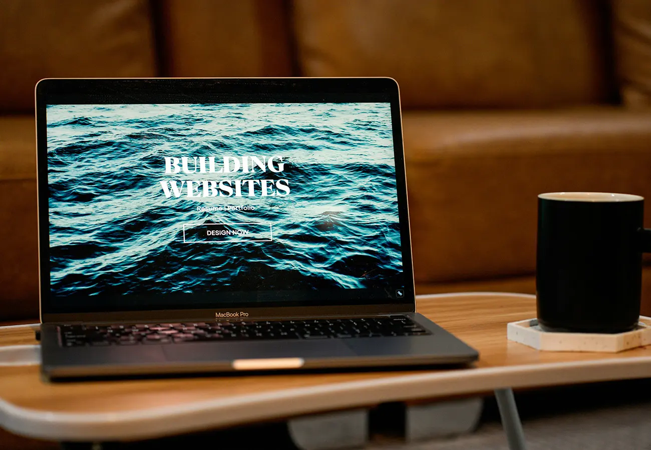 MacBook Pro on a wooden table showing a 'BUILDING WEBSITES' webpage with a 'DESIGN NOW' button and a black mug nearby on a coaster